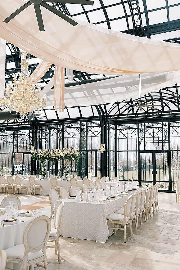 Reception tablescape with a long banquet table wedding setup, white linens and greenery garland under a chandelier in a glass conservatory.