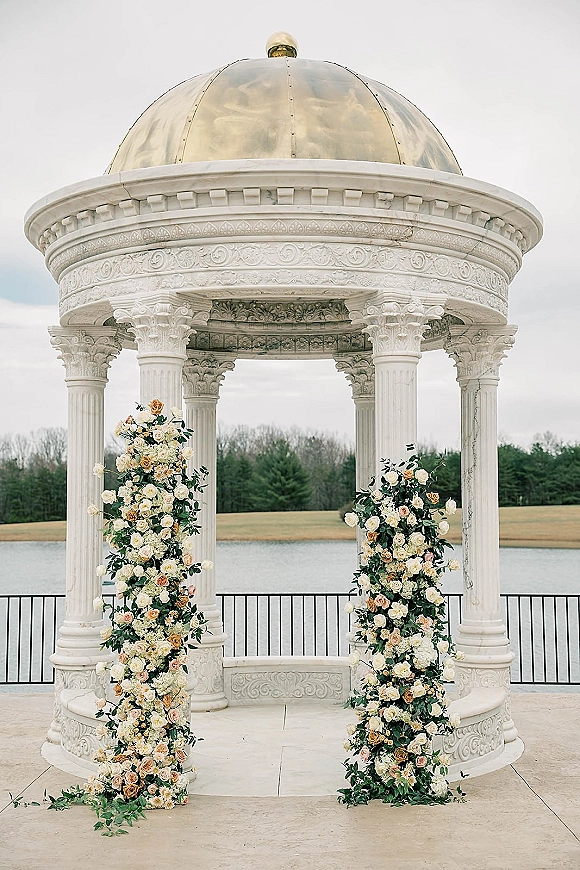 Wedding ceremony backdrop with twin floral pillars of white and blush roses on marble columns beneath a domed lakeside gazebo