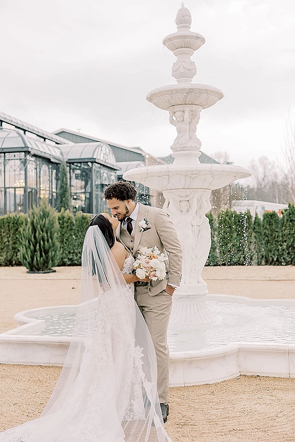 Wedding kiss portrait of bride and groom kissing, her long veil and bouquet beside a stone fountain in a greenhouse courtyard under overcast sky