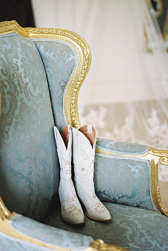 Wedding boots in white cowboy boots wedding style with embroidery and gold trim, resting on a vintage upholstered chair indoors