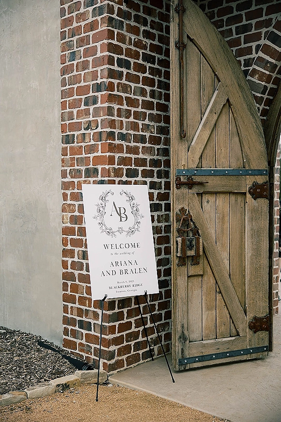 Wedding welcome sign with a monogram crest and floral wreath illustration on an easel by a brick wall and rustic wooden door entrance