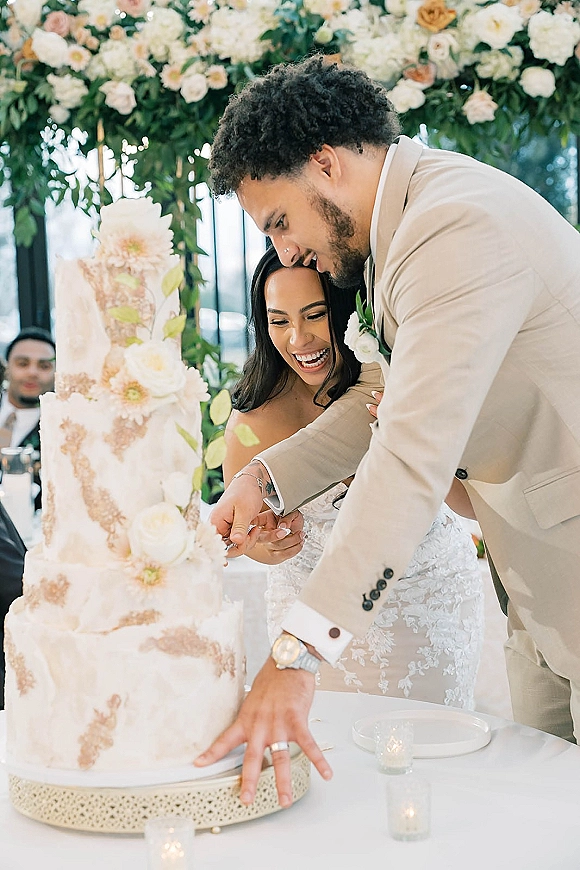 Cake cutting by bride and groom as they slice a tall white wedding cake with fresh flowers on a candlelit reception table before guests