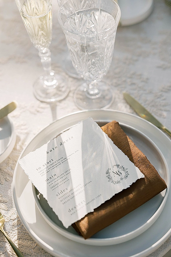 Wedding place setting with a wedding menu card on deckle edge paper, monogram crest and gold flatware on a lace tablecloth