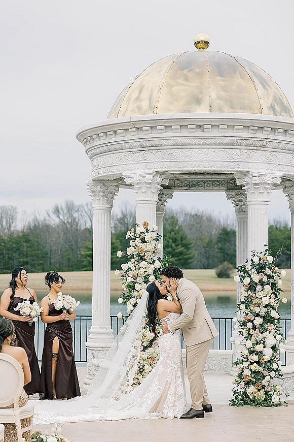 Ceremony kiss at an outdoor wedding ceremony under a floral arch, bride in lace dress and veil kissing groom by a lakeside gazebo
