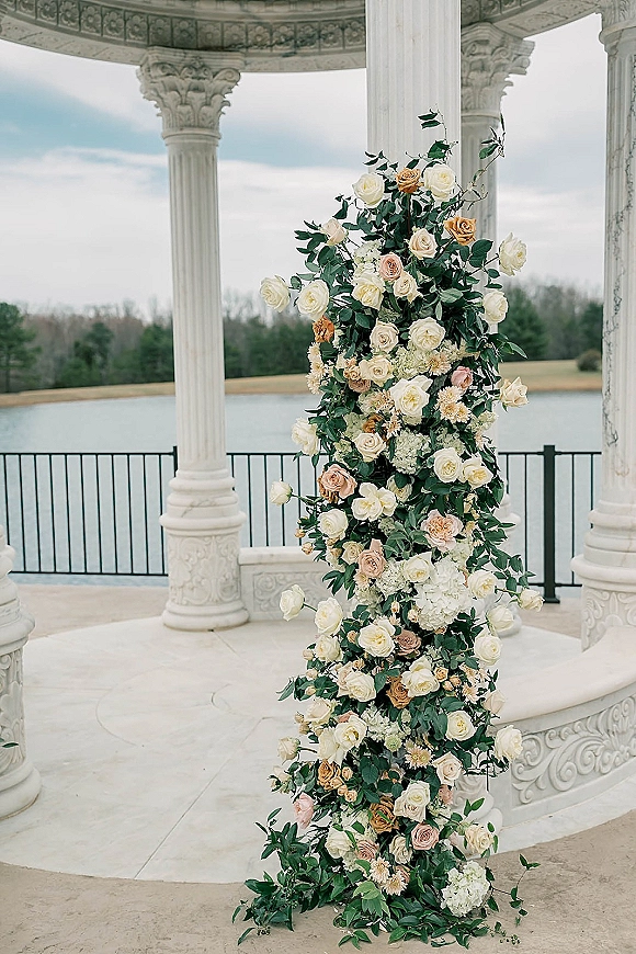 Wedding ceremony flowers with ceremony floral pillar of roses, hydrangea, and greenery framing gazebo columns by a lakeside pavilion