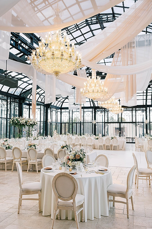 Wedding reception decor with crystal chandeliers above draped ceiling, round white-linen tables and floral centerpieces in a glass conservatory setting