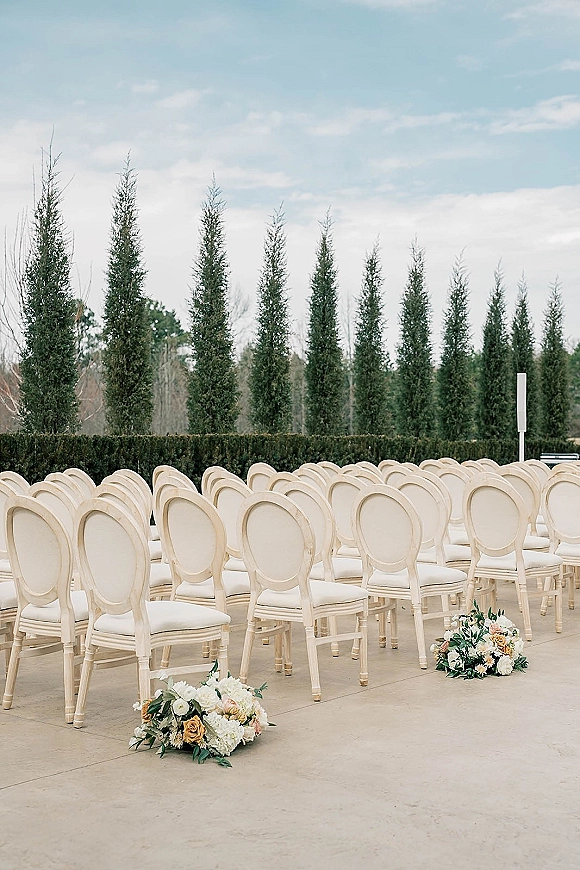 Ceremony seating setup with outdoor wedding ceremony chairs in neat rows, white chairs lining a patio aisle with peach floral clusters and greenery by hedges