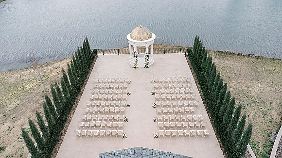Ceremony setup for an outdoor wedding ceremony with white chairs facing a gazebo altar, floral columns lining the aisle by a lake