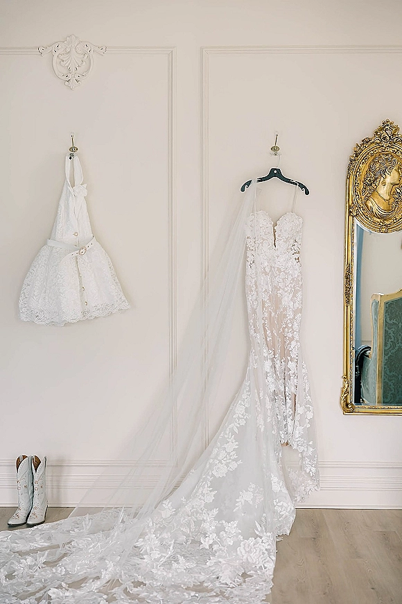 Wedding dress with lace wedding dress detailing hanging beside an ornate gold mirror, long veil trailing over wood floor by white paneled wall
