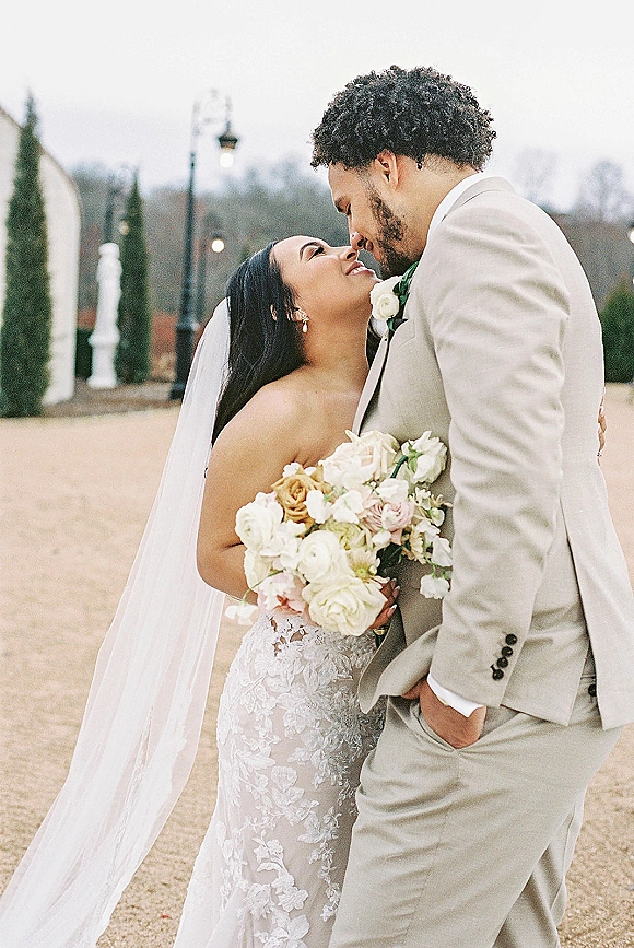 Wedding couple portrait of bride and groom embrace, touching foreheads as she holds a white rose bouquet on an overcast lawn with trees