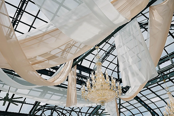 Ceiling draping decor with sheer fabric swags and a crystal chandelier beneath a glass skylight ceiling with black metal beams