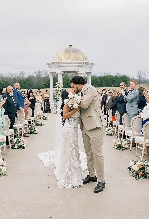 Ceremony kiss as bride holds bouquet and kisses groom in beige suit beneath a floral arch in an outdoor garden rotunda with guests cheering