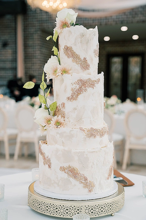Wedding cake with white frosting and gold lace appliques, sugar flowers, and greenery vine on a stand in a brick-walled reception room