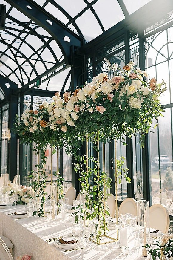 Reception tablescape with wedding table centerpiece of roses and hydrangea on gold stands, taper candles on a white linen table in a sunny glass conservatory