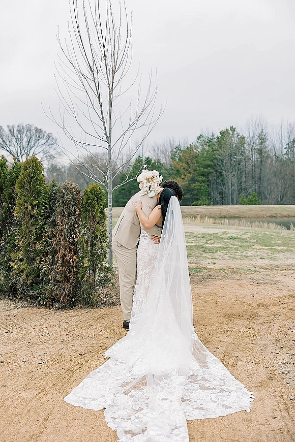 Wedding couple portrait of bride and groom hugging, her long veil and lace dress with bouquet over shoulder on a gravel path in a field
