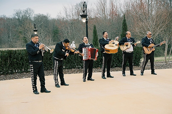 Wedding mariachi band in matching charro suits with trumpet, violin, accordion and guitars posing in an outdoor courtyard under overcast skies