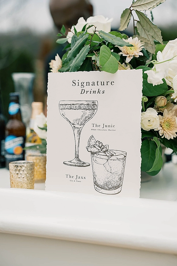 Signature drinks sign with wedding signature drinks menu on deckle-edge card, cocktail sketches, and floral greenery on a bar counter