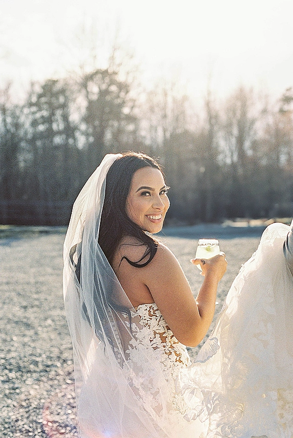 Bridal portrait of a bride looking back in a strapless lace dress with long veil, holding a cocktail glass in a sunlit field