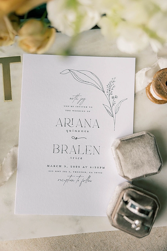 Wedding invitation with minimalist wedding invitation typography, botanical line art, wax seal and ribbon beside ring box on light fabric with blurred flowers