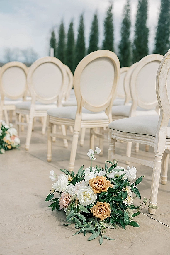 Ceremony aisle decor with aisle floral arrangements of roses and greenery lining a stone patio between cream oval-back chairs outdoors