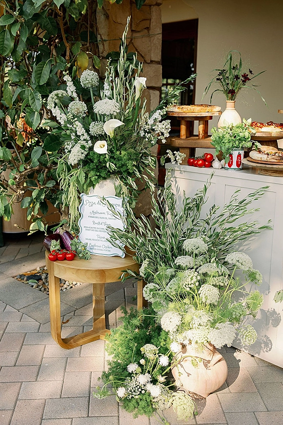 Pizza station with wedding pizza station menu sign, tiered pies on stands, and calla lily greenery accents on a patio by a stone wall