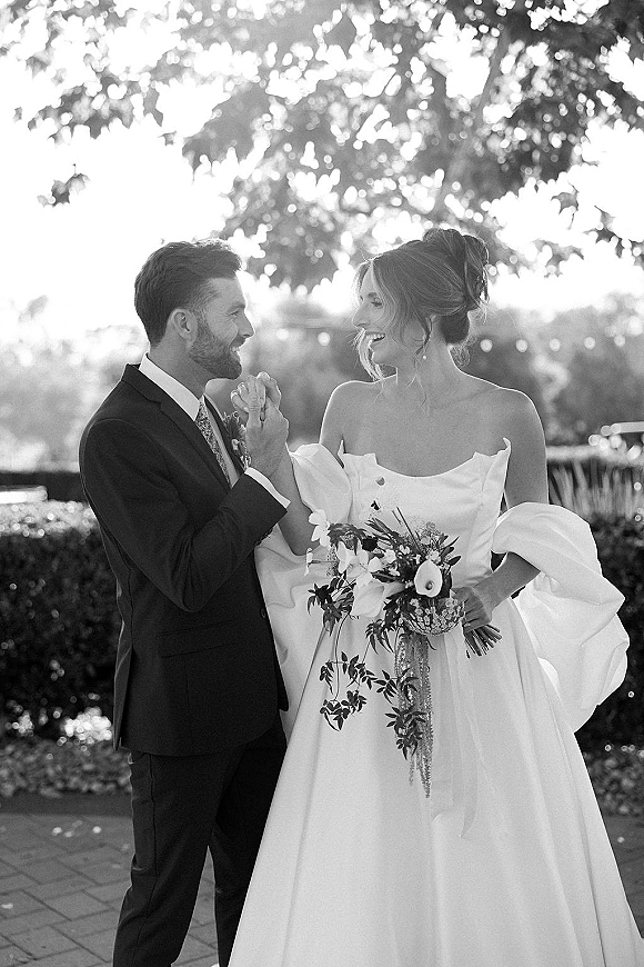 Couple portrait of bride and groom holding hands, bride laughing in strapless gown with puff sleeves and bouquet in sunlit garden greenery