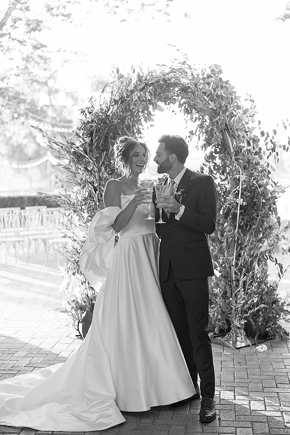 Wedding couple portrait of newlyweds toasting with champagne flutes under a greenery arch with string lights on an outdoor brick patio