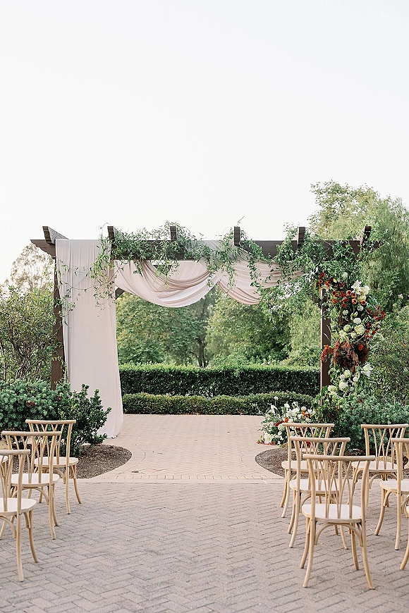 Ceremony setup with wood pergola draped in fabric, greenery garland and asymmetrical florals lining a stone paver aisle in a hedge garden courtyard