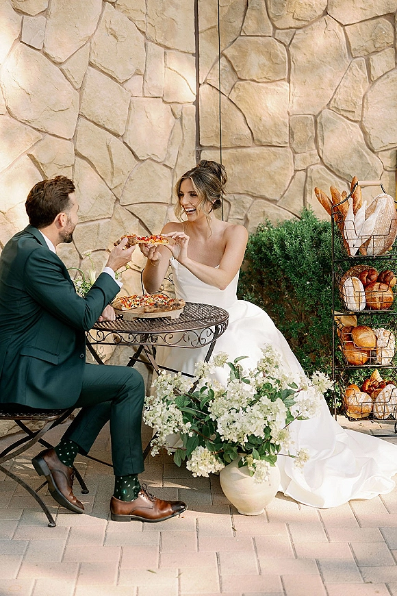 Couple portrait of bride and groom eating pizza at a bistro table, bride in strapless dress and groom in green suit by a stone wall
