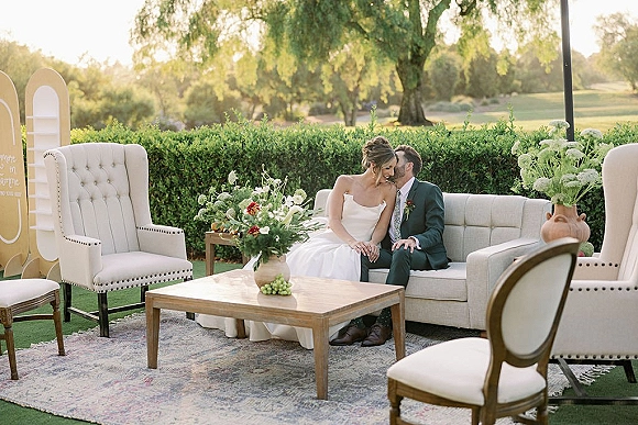 Couple portrait of bride and groom on sofa in an outdoor wedding lounge, with wedding gown and green suit beside white florals on a coffee table