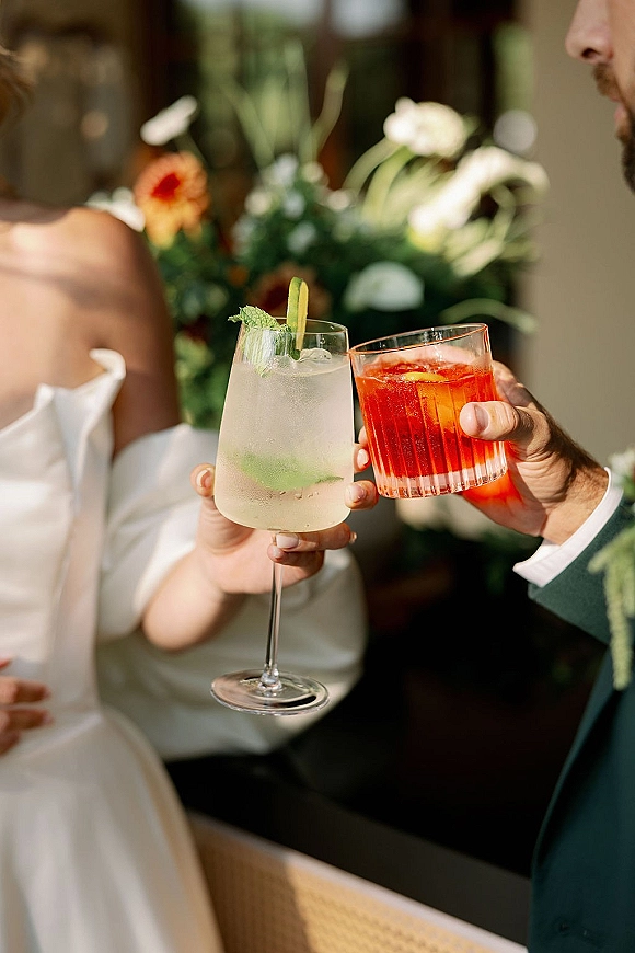 Wedding toast as newlyweds clinking glasses with mint and citrus garnishes, bride in strapless gown and groom boutonniere amid greenery
