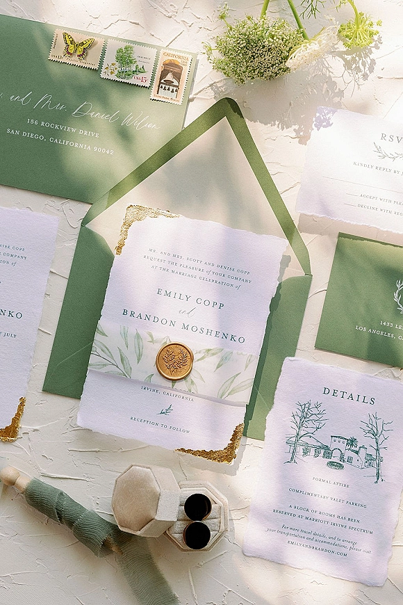 Invitation suite with green wedding invitations, deckle edge paper, gold wax seal, and floral sprigs on a textured white surface