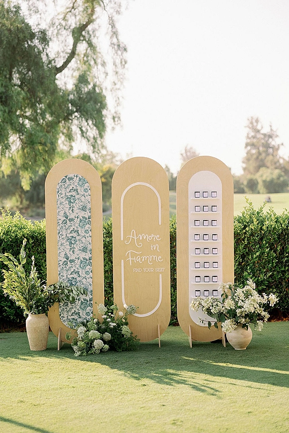 Wedding seating chart escort card display on arched panels with calligraphy sign, greenery garland, and floral vases on a garden lawn