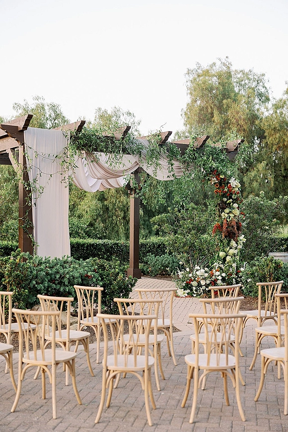 Ceremony setup with a wood pergola altar, draped fabric and hanging greenery, lined with wooden chairs on a brick paver patio in a garden setting