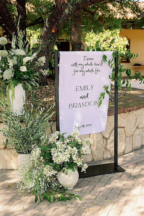 Wedding welcome sign on linen fabric with calligraphy lettering, framed by white flowers and greenery on a black metal stand by trees and stone wall