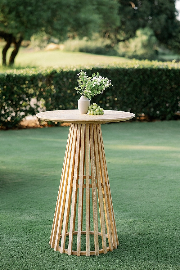 Cocktail table decor with a white vase of white flowers and greenery, plus green grapes on a wood high-top table on a lawn with hedges