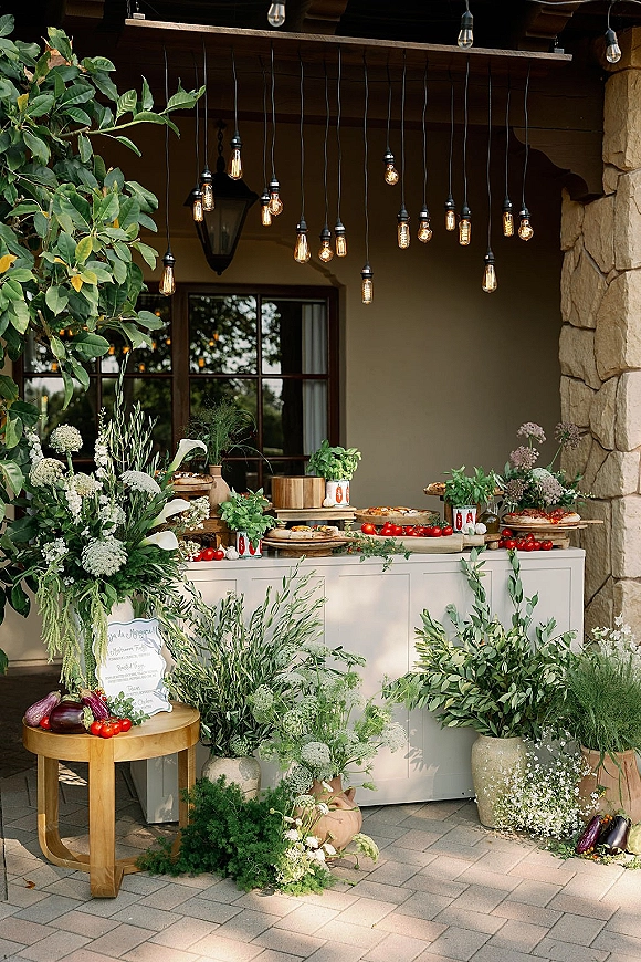 Wedding food station with pizza wedding catering on a white buffet counter, topped with greenery garland and Edison bulbs on a patio