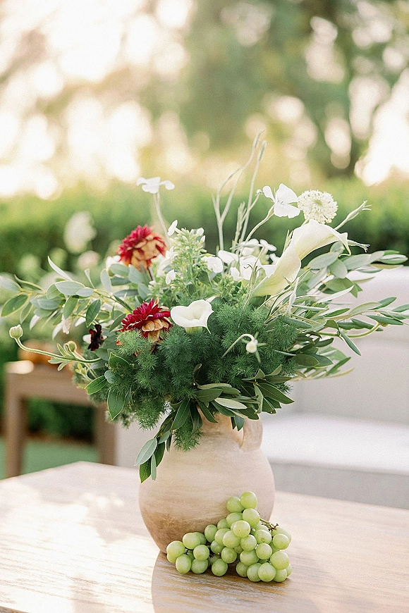 Wedding centerpiece with floral centerpiece idea of calla lilies and red-white blooms in a ceramic vase, set on a wooden table outdoors