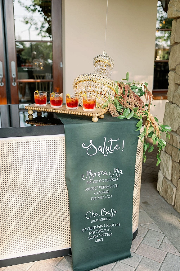 Wedding bar setup with a cocktail menu sign, champagne coupe tower, and lemon-garnished drinks on a rattan front bar on a patio