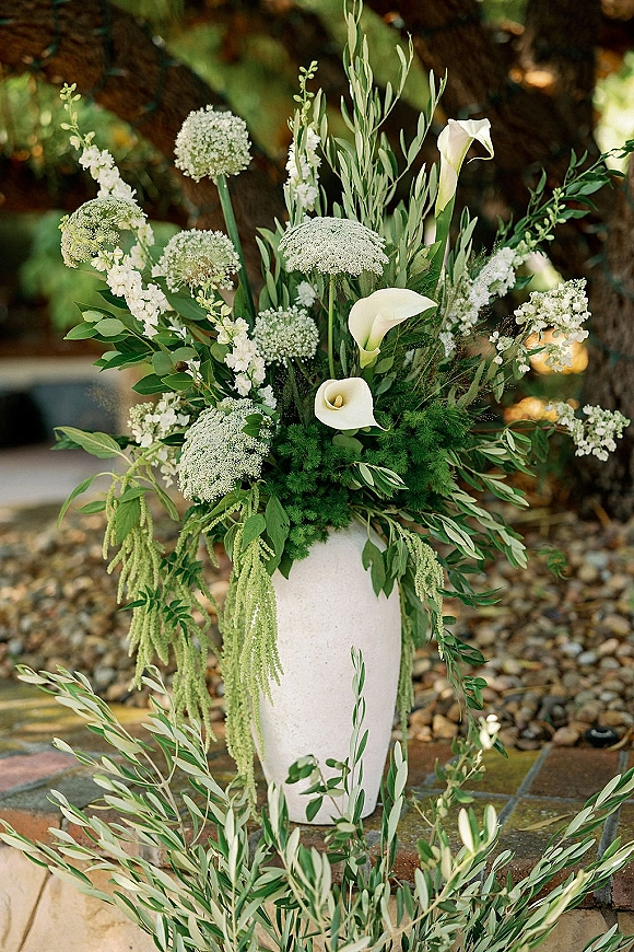 Wedding floral arrangement with ceremony aisle flowers in a tall white vase, featuring calla lilies, white blossoms, and olive greenery by stone steps outdoors