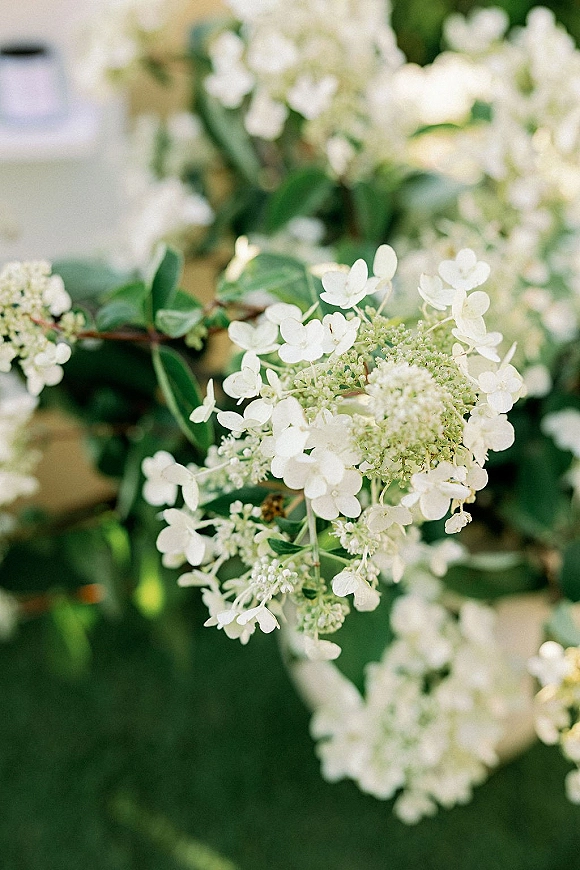 Wedding florals with white wedding flowers and lush greenery arranged in a full centerpiece, with blurred garden foliage in the background