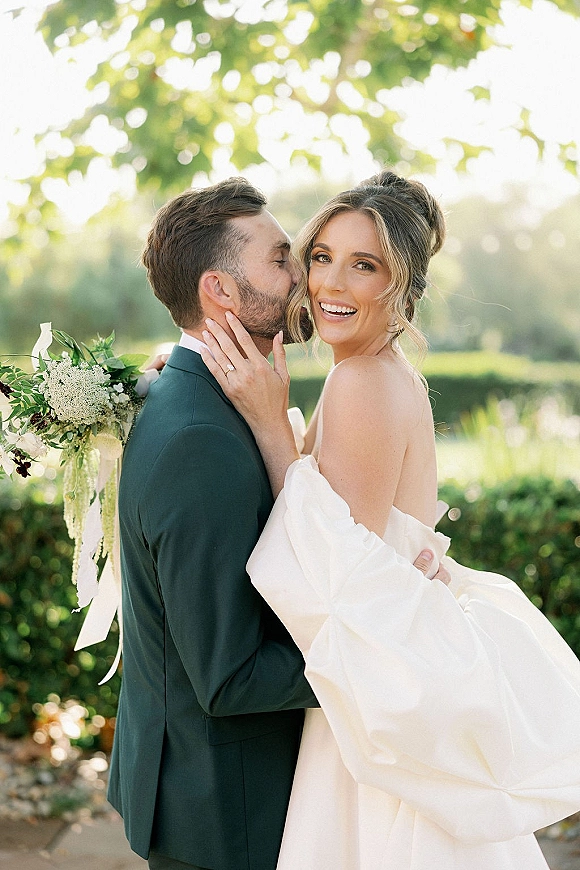 Wedding couple portrait of groom kissing bride’s cheek as she looks at camera, holding a white-green bouquet with ribbon by a sunny pond