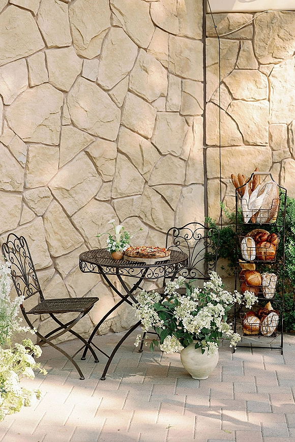 Wedding cocktail hour decor with a wedding bread display of baguettes and loaves beside bistro table and chairs on a brick patio by a stone wall