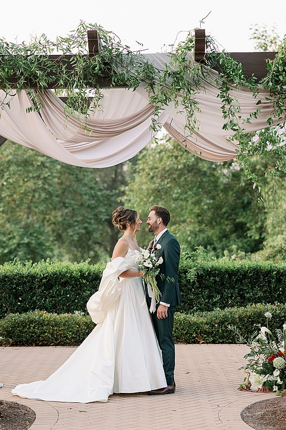 Ceremony couple portrait of bride and groom under arch, forehead touching, with bouquet and greenery garland on a brick walkway in garden hedges
