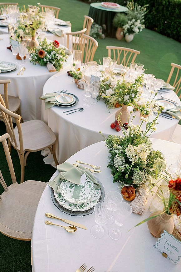 Reception tablescape with an outdoor reception table featuring white linens, floral centerpieces, gold flatware, candles, and place cards on a green lawn