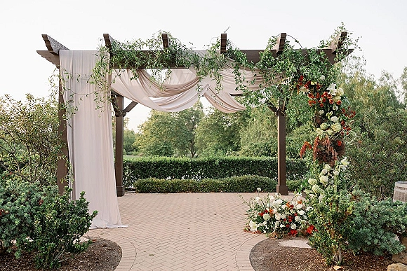 Wedding ceremony arch with draped fabric and greenery garland, accented by red and white florals on a garden patio with trees