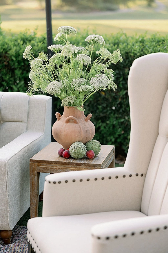Wedding lounge decor with upholstered armchairs and a wooden side table, featuring a terracotta vase of white wildflowers against hedge greenery.