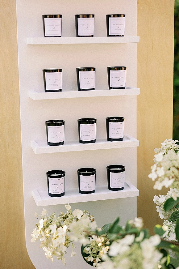 Wedding escort display of black candles with printed name labels on white shelves, accented by floral arrangements and greenery against a wooden wall
