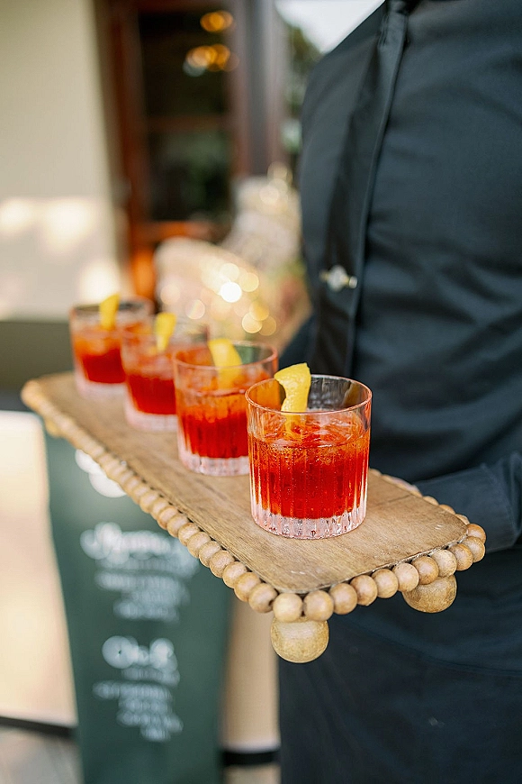 Signature cocktails in rocks glasses with citrus peel garnish on a wooden serving tray, carried by a server on an outdoor patio with chalkboard sign