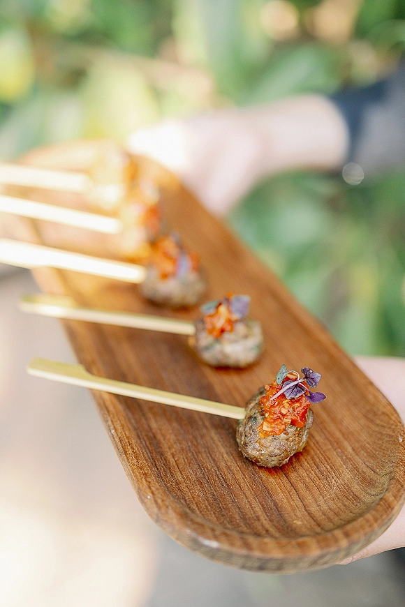 Wedding cocktail appetizers on a wood serving board with passed hors d’oeuvres skewers and microgreens, held up against blurred outdoor greenery